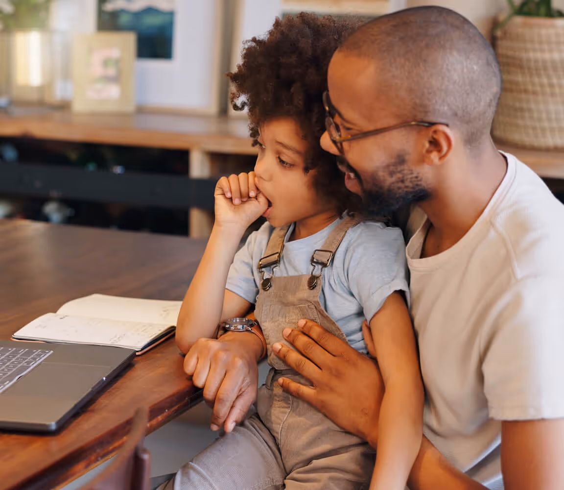 Man with glasses sitting with a child who is sucking their thumb, both looking at a laptop on a wooden table.