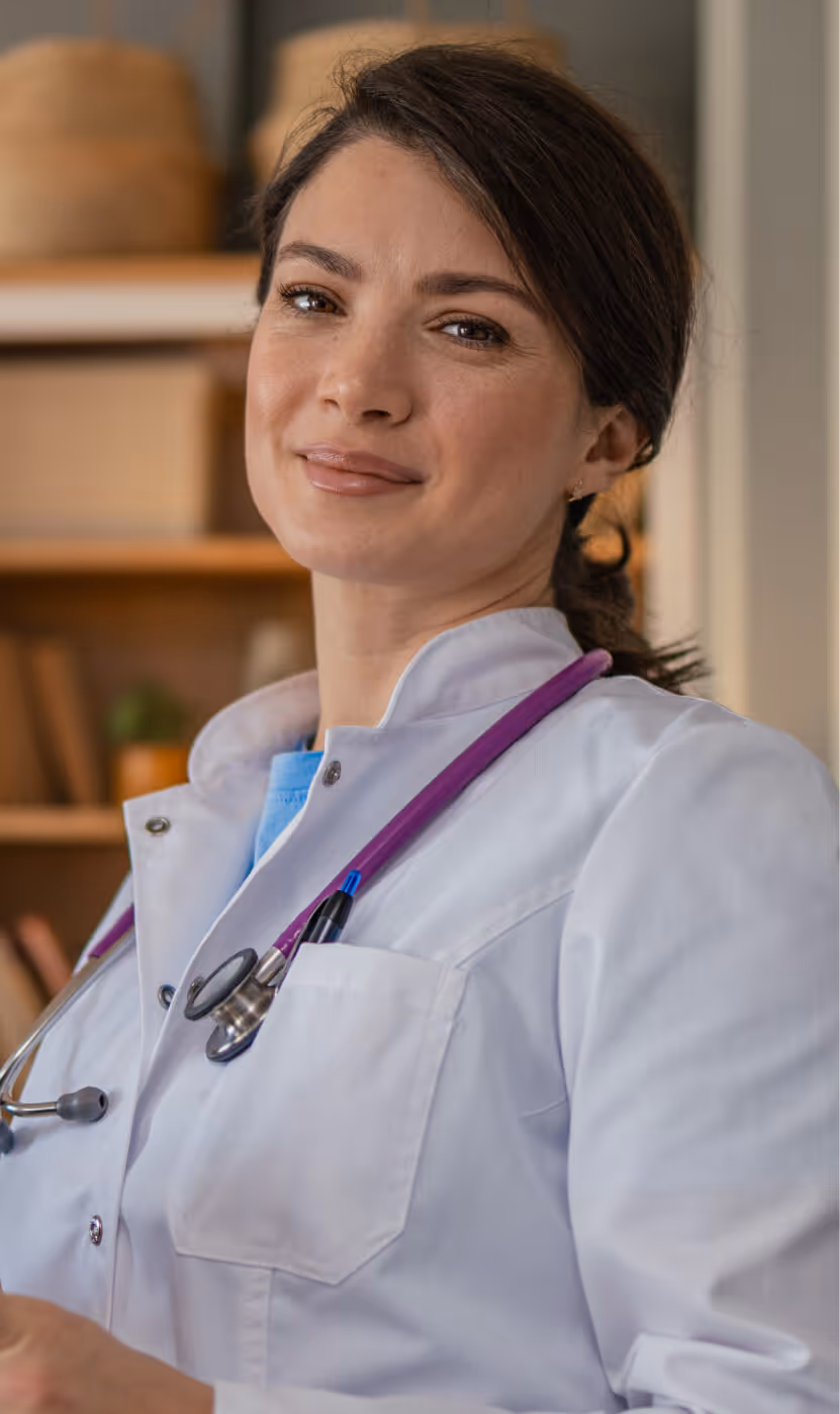 Smiling female doctor wearing a white coat with a purple stethoscope around her neck.