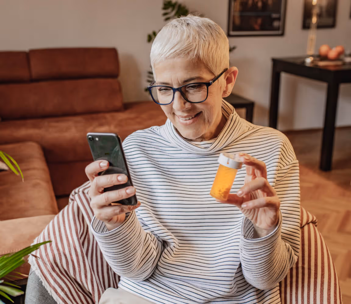 Smiling elderly woman sitting indoors holding a prescription pill bottle in one hand and a smartphone in the other.