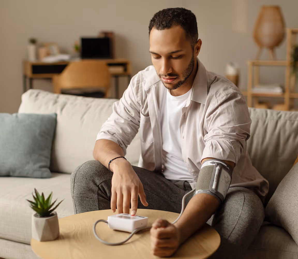 Man sitting on a couch checking his blood pressure with an automatic monitor on a wooden table.