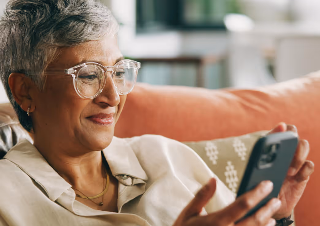 Older woman with short gray hair and clear glasses smiling while looking at her smartphone on a couch.