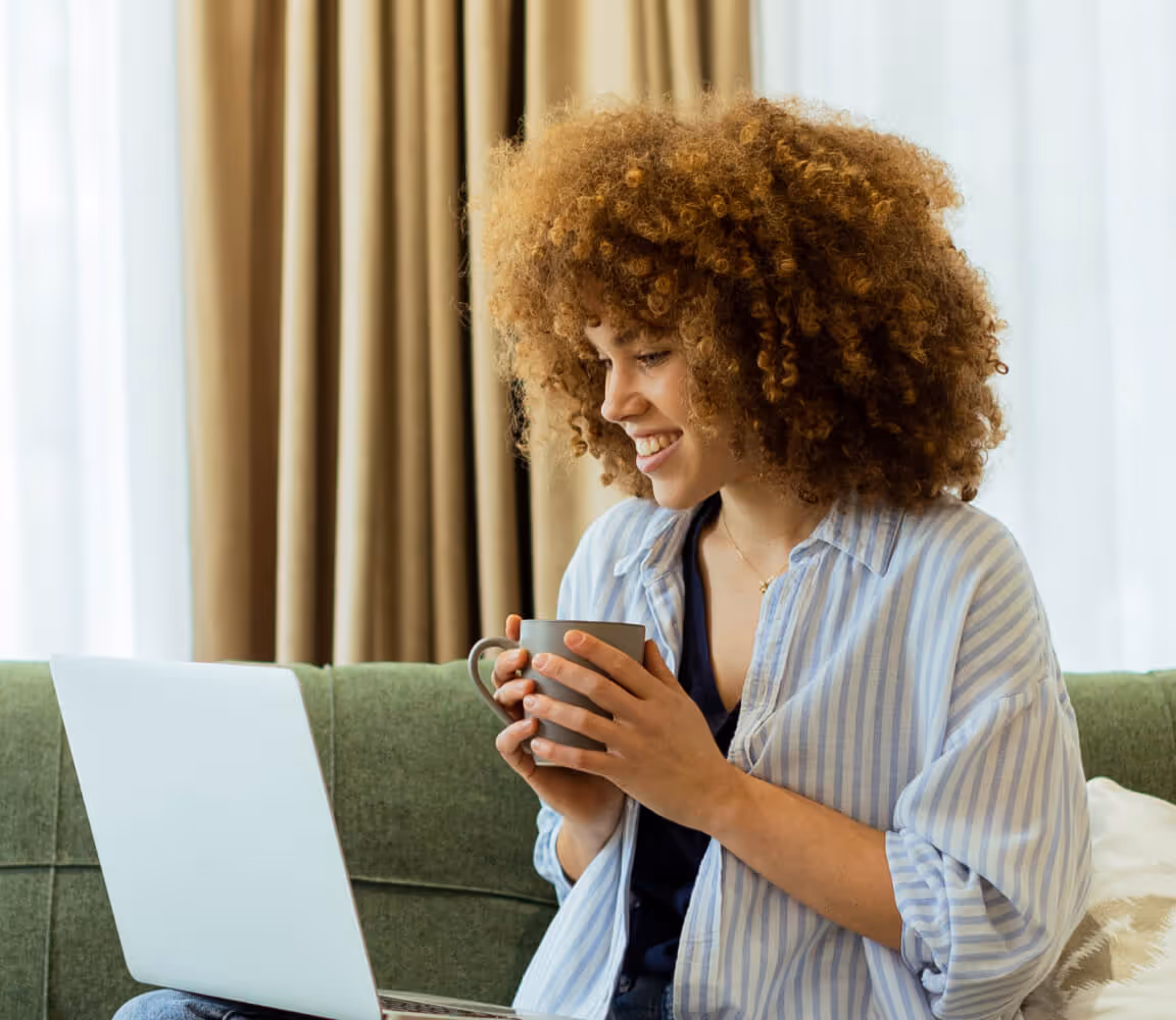 Smiling woman with curly hair sitting on a couch holding a mug and looking at a laptop.