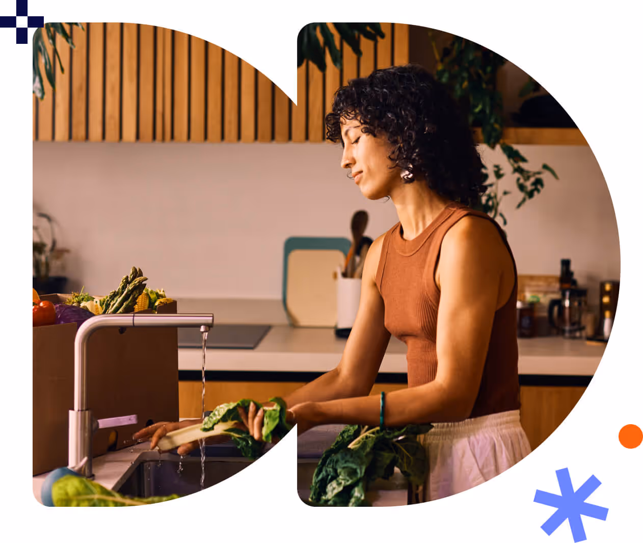 Woman washing leafy greens at a kitchen sink next to a box of fresh vegetables.
