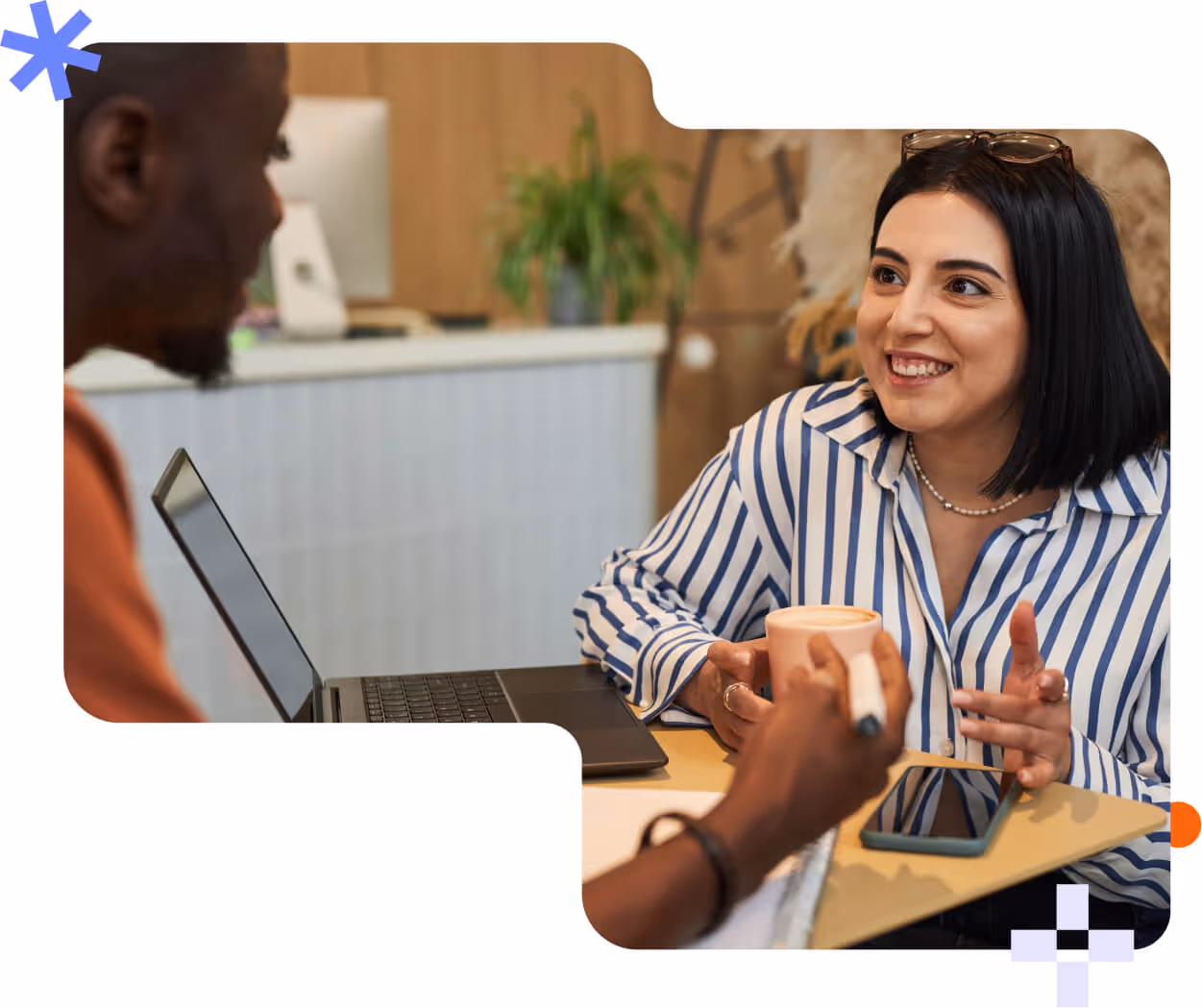 Two colleagues having a friendly conversation over a coffee in an office setting with a laptop and smartphone on the table.