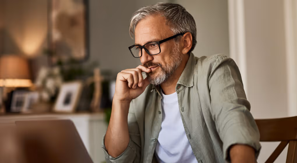 Middle-aged man with gray hair and glasses thoughtfully looking at a laptop screen indoors.