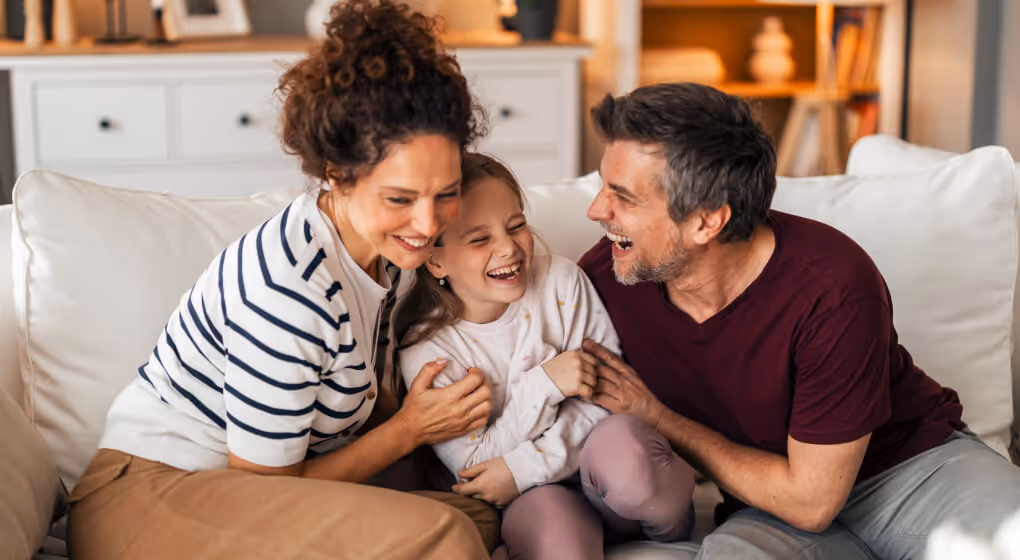 Smiling parents sitting on a couch laughing and hugging their happy young daughter in a cozy living room.