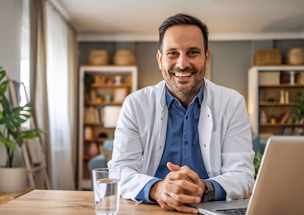 Smiling male doctor in white coat sitting at a desk with a laptop and glass of water in a bright office.
