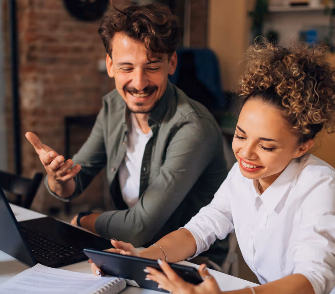 Two colleagues smiling and discussing while looking at a tablet and laptop in a casual office setting.