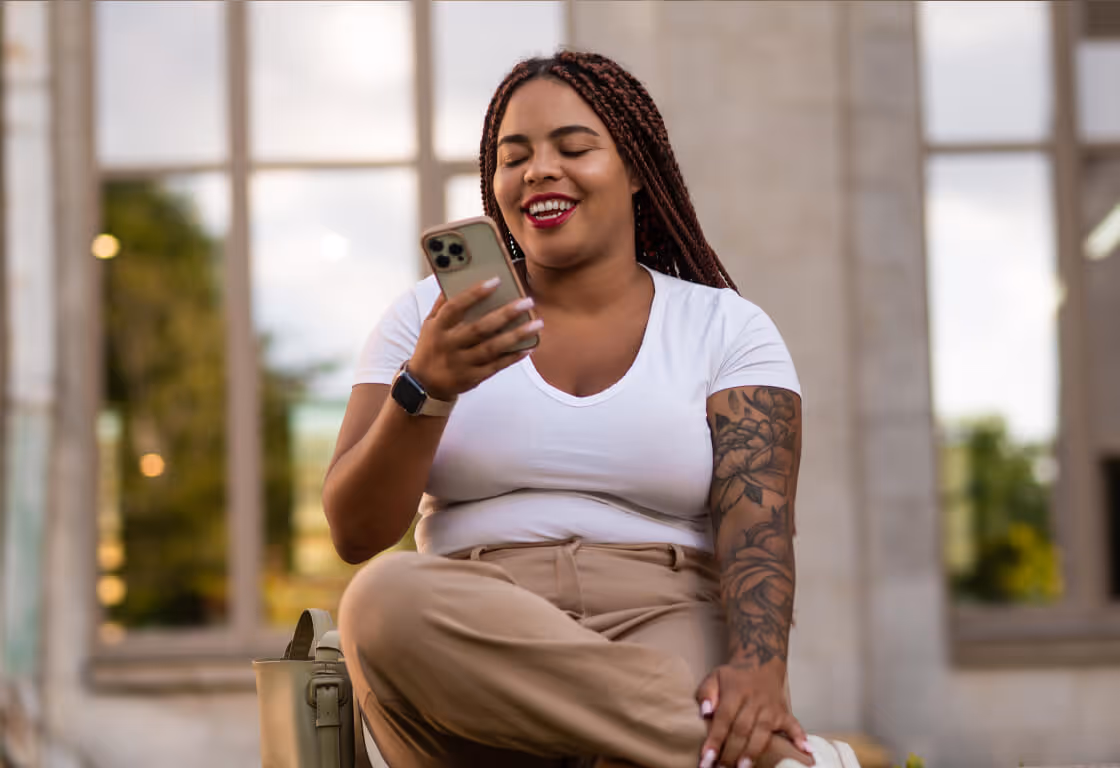Smiling woman with braided hair and tattoo on her arm sitting cross-legged outdoors, looking at her smartphone.