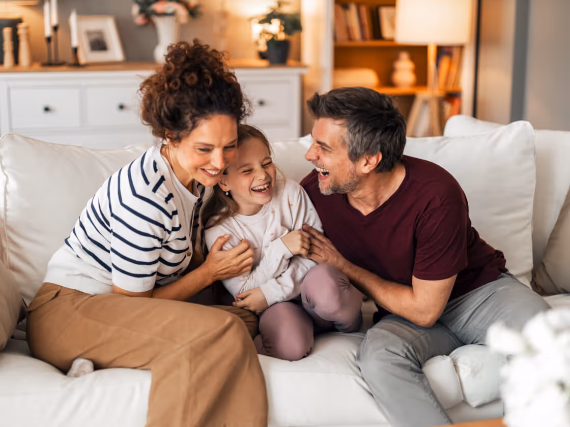 A joyful family of three laughing together on a white couch in a cozy living room.