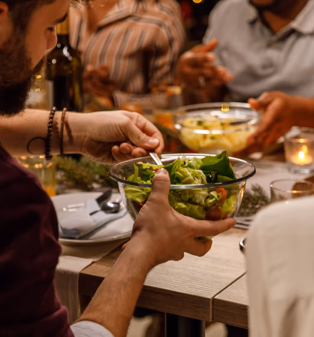 Person mixing fresh green salad in a glass bowl at a dinner table with others.