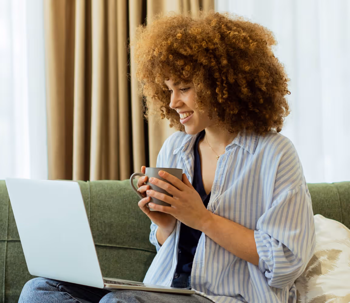Smiling woman with curly hair sitting on a couch, holding a mug and looking at a laptop.