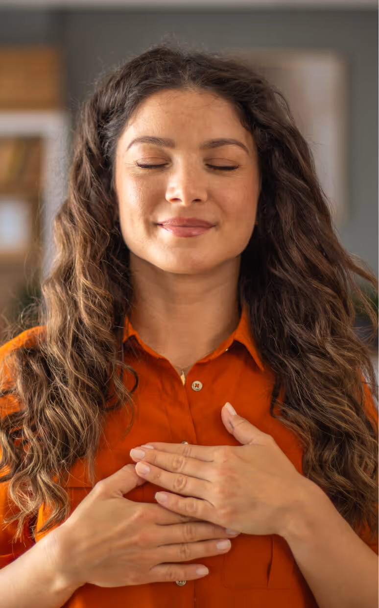 Woman with closed eyes and long curly hair wearing an orange shirt, placing her hands on her chest in a calm, reflective pose.