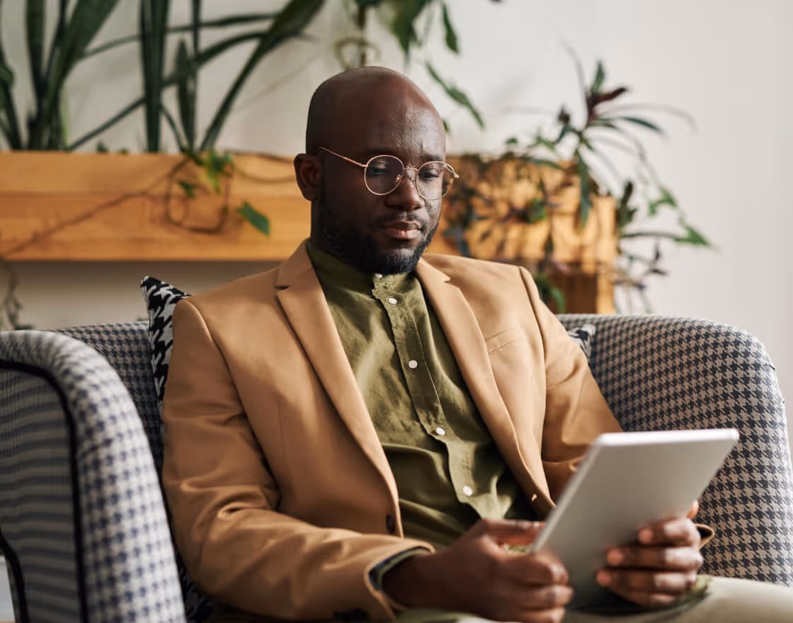 Man wearing glasses and a beige blazer sitting on a patterned armchair, reading a tablet.