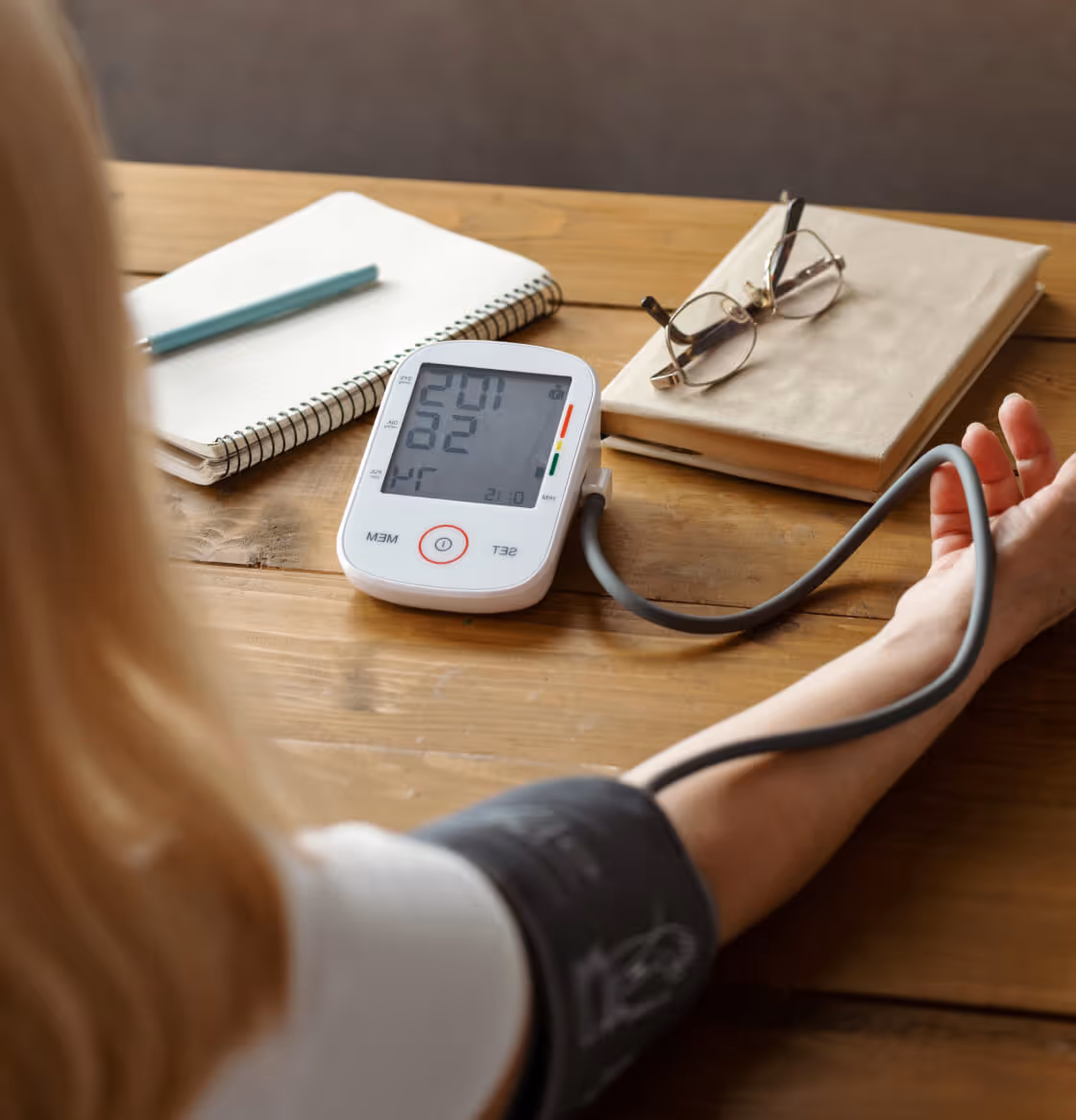 Person measuring their blood pressure with a digital monitor on a wooden table next to notebooks and eyeglasses.