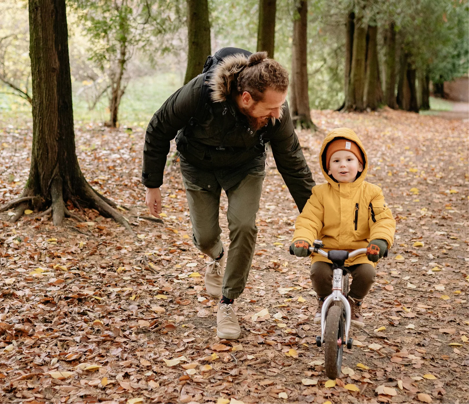 A father walks beside his young child on a balance bike in an autumn park.
