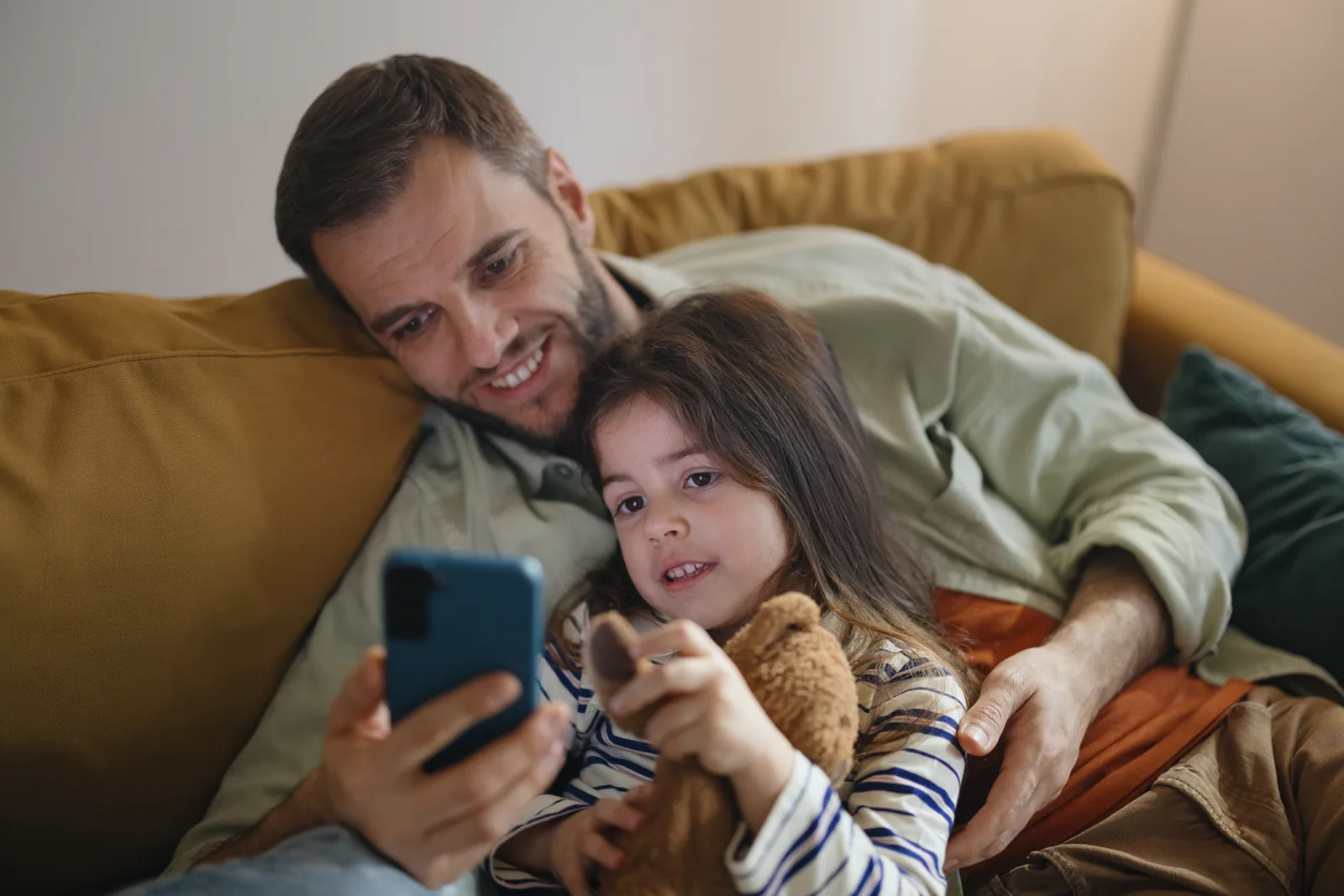 A father and daughter look at a phone together on the couch.