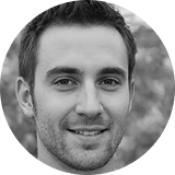 Smiling young man with short dark hair and light stubble, outdoors background.