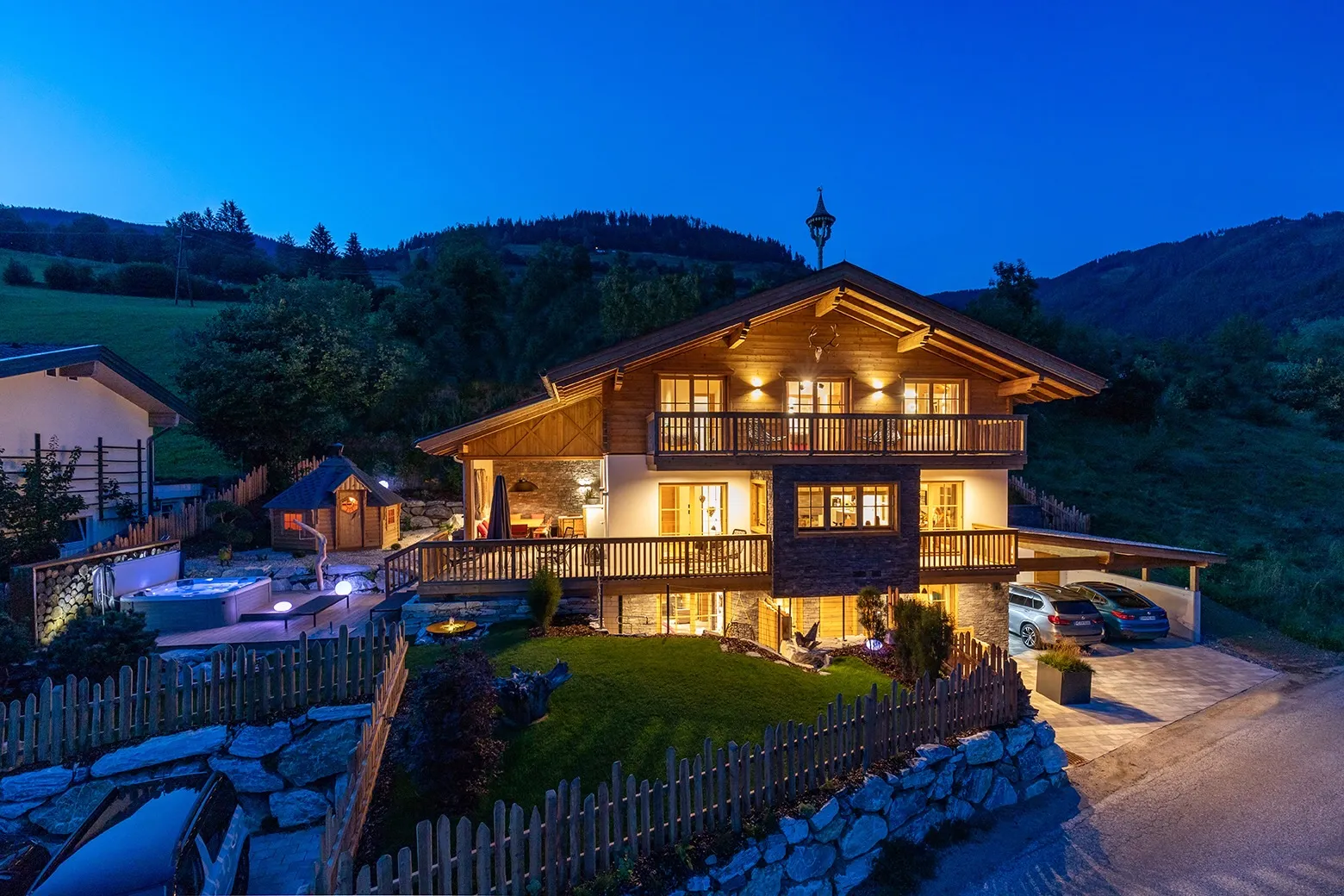 Illuminated wooden chalet with balconies, a fenced garden, hot tub, and parked cars at dusk in a mountainous area.