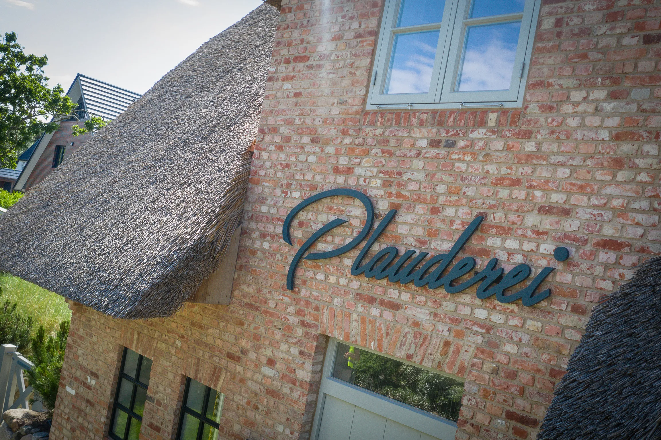 Brick building with thatched roof and blue sign reading 'Plauderei' under a window reflecting the sky.