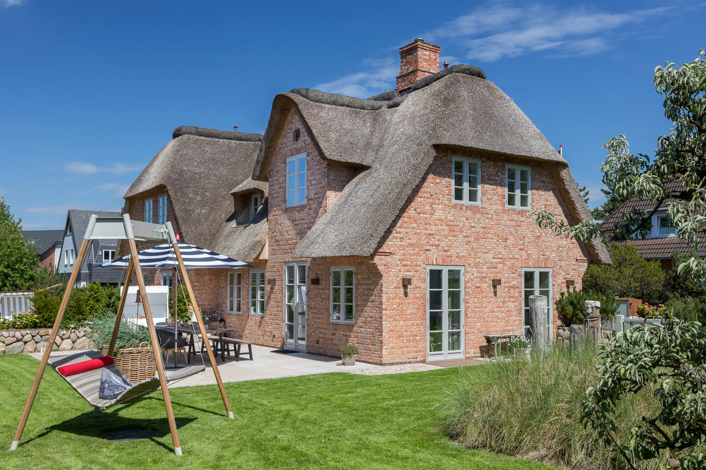 Modern brick house with a thatched roof, patio with outdoor furniture, and a hammock swing on a green lawn under a clear blue sky.