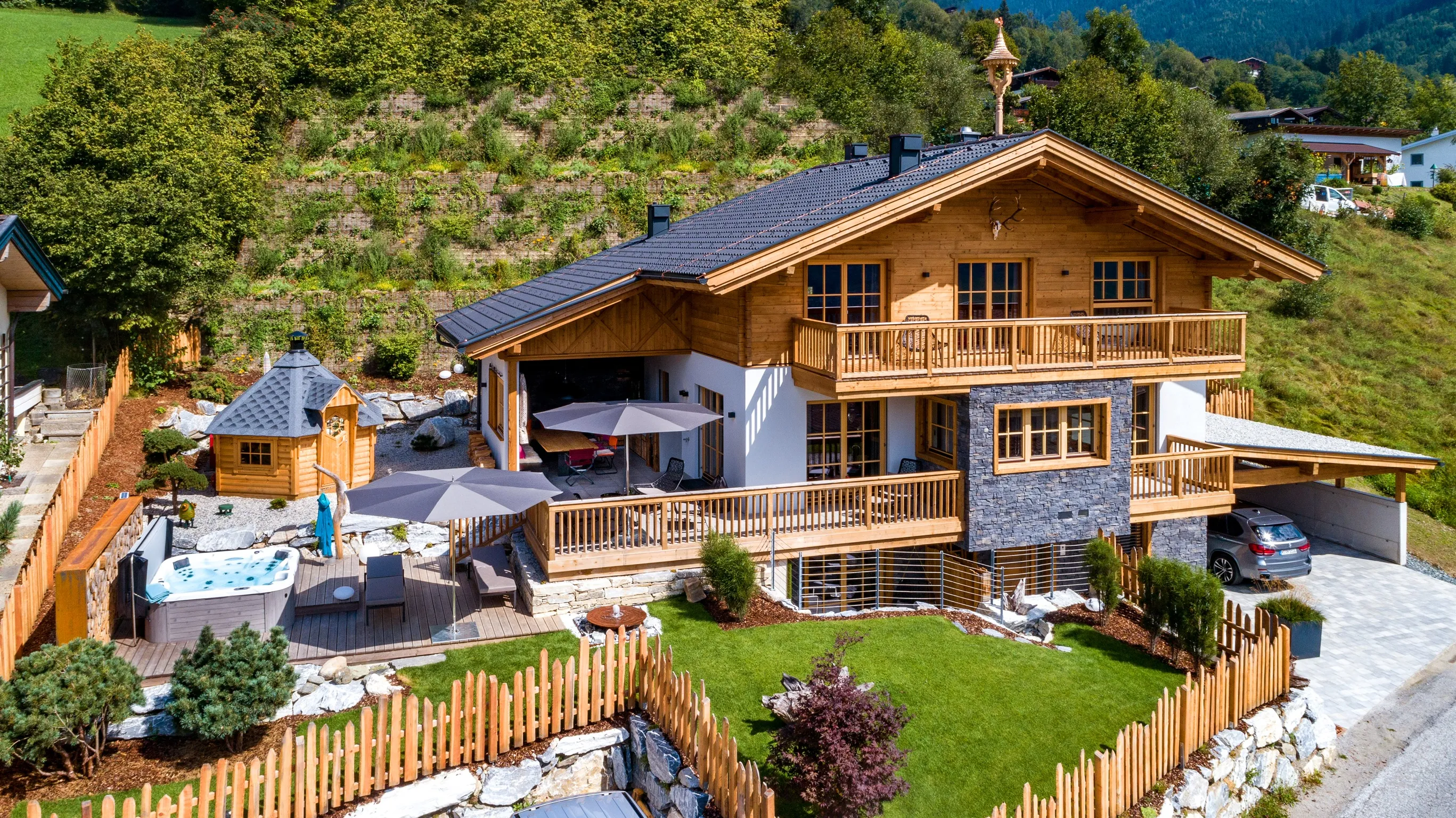 Modern wooden house with balconies, a fenced green lawn, outdoor seating, umbrellas, a hot tub, and a car parked in a covered driveway.