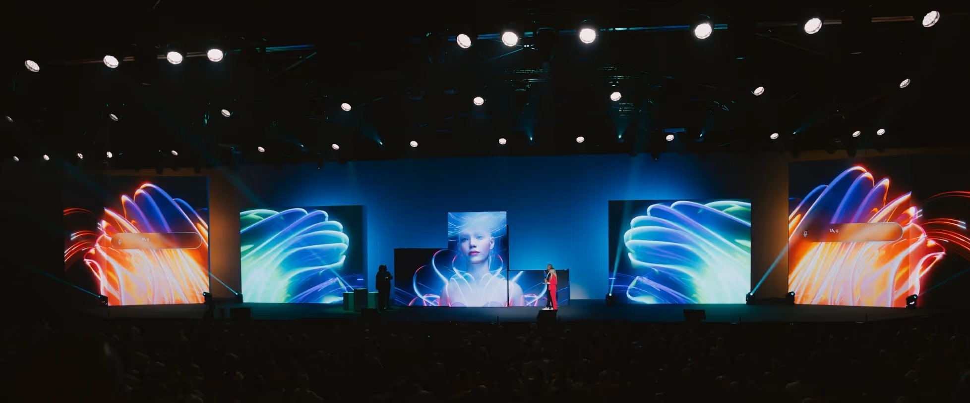 Stage with large digital screens displaying colorful abstract shapes and a woman's face, with two people standing on the stage under bright spotlights.