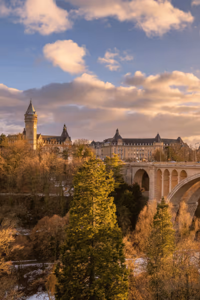 Stone arch bridge over trees with large historic building and cloudy sky at sunset.