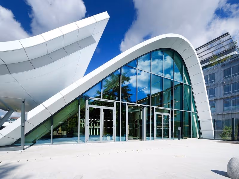 Modern building with curved white roof and large glass windows reflecting a blue sky with clouds.