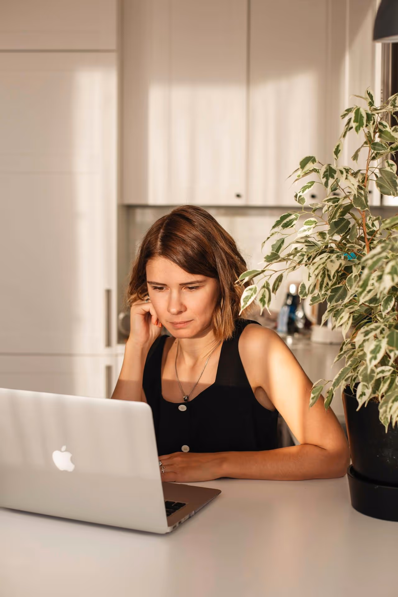 Woman in black sleeveless top focused on a laptop, seated at a white table with a leafy plant nearby.