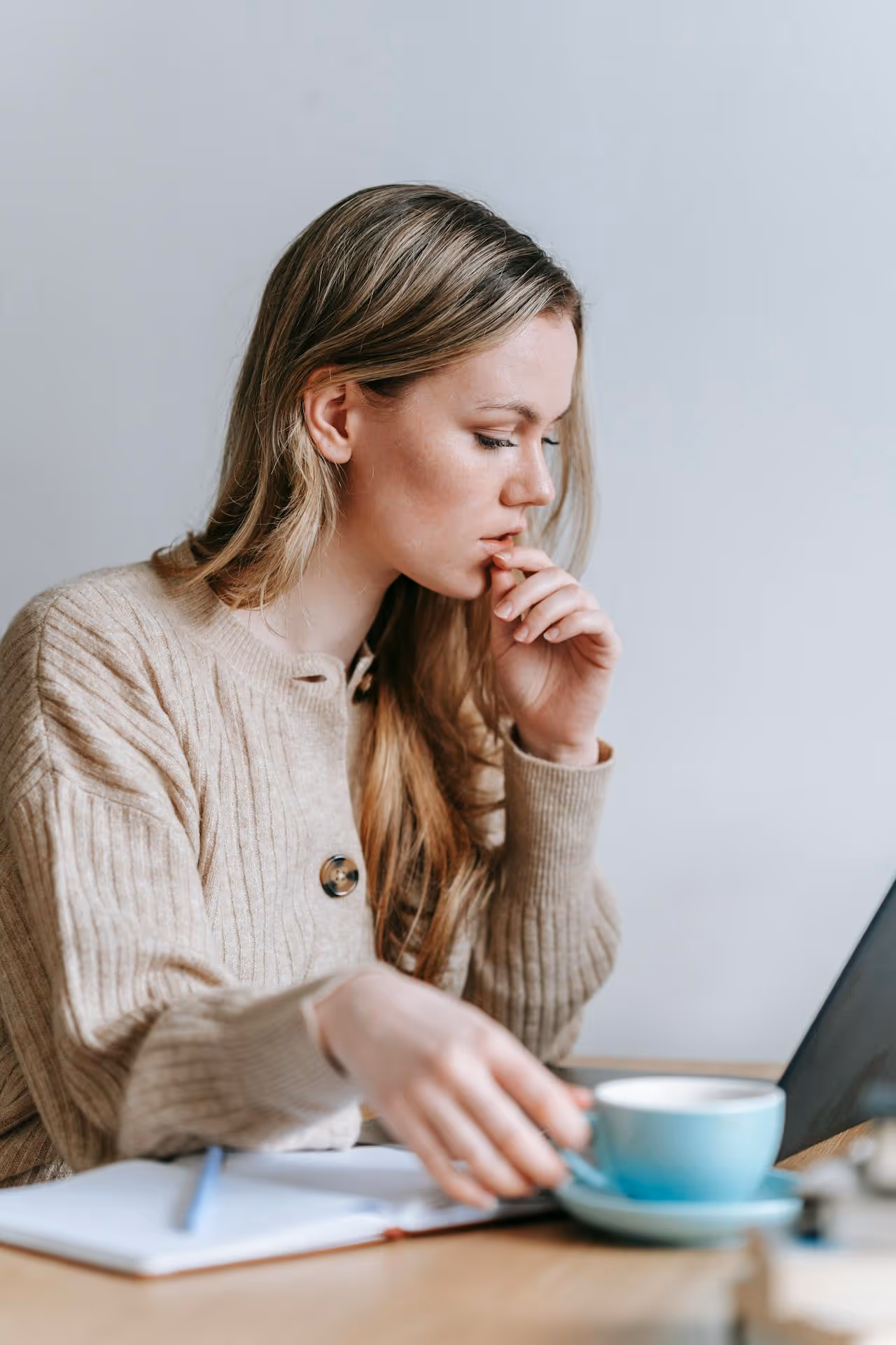 Focused woman in a beige sweater holding a blue coffee cup while looking at a laptop screen.