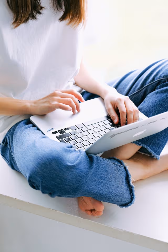 Person in white shirt and jeans sitting cross-legged using a laptop on their lap.