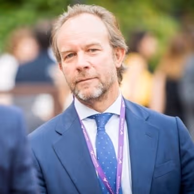 Man with beard and blue suit standing outdoors with blurred people and greenery in the background.