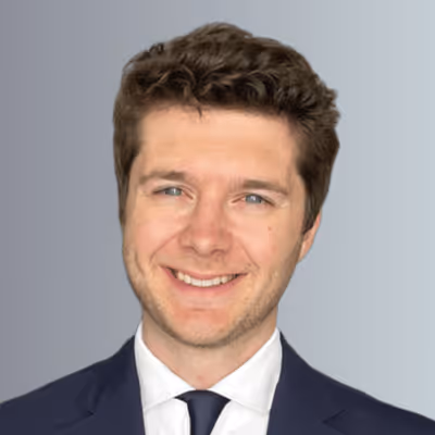 Smiling young man with curly brown hair wearing a dark suit, white shirt, and black tie on gray background.
