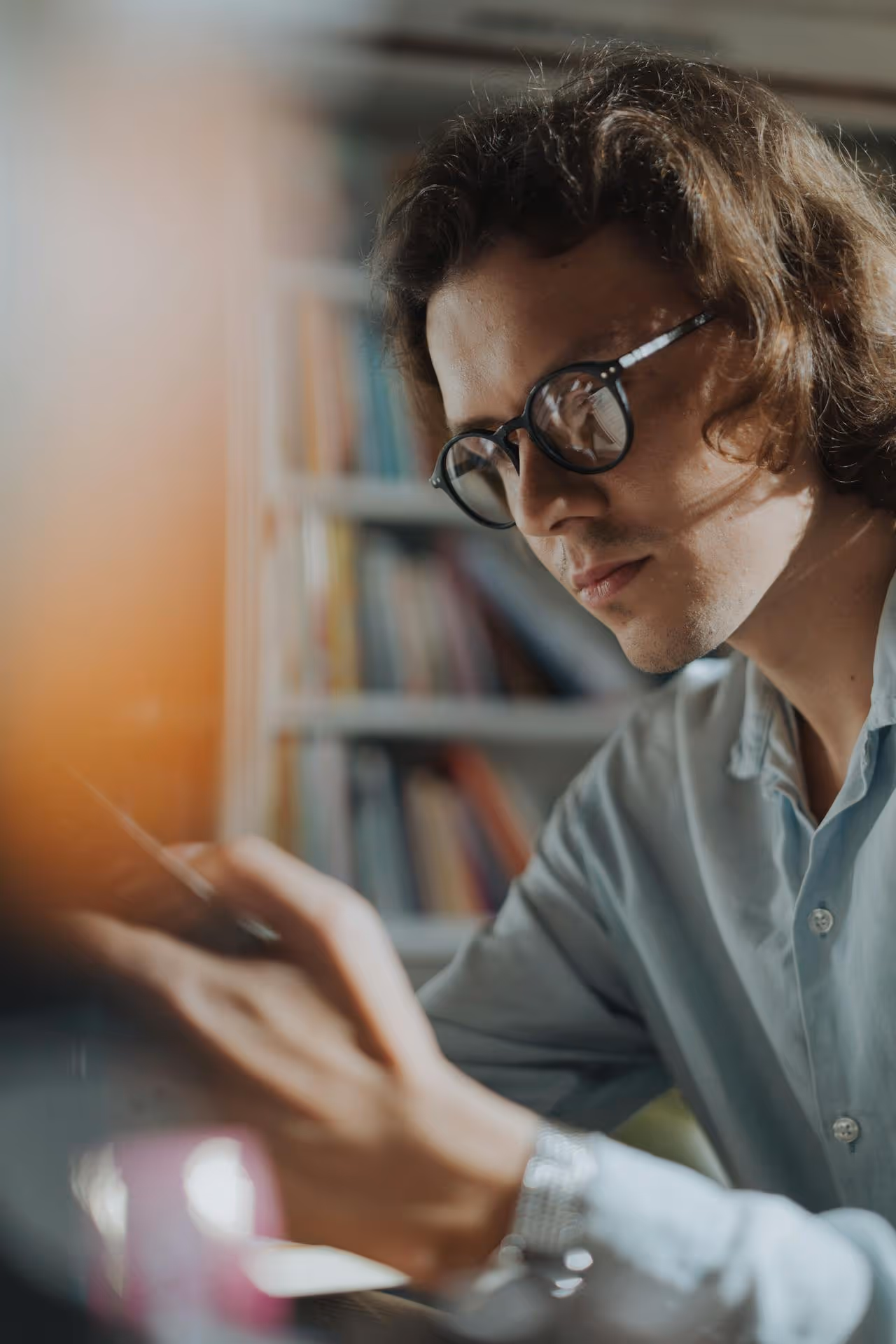 Young man with glasses focused on writing or drawing at a desk with a bookshelf in the background.