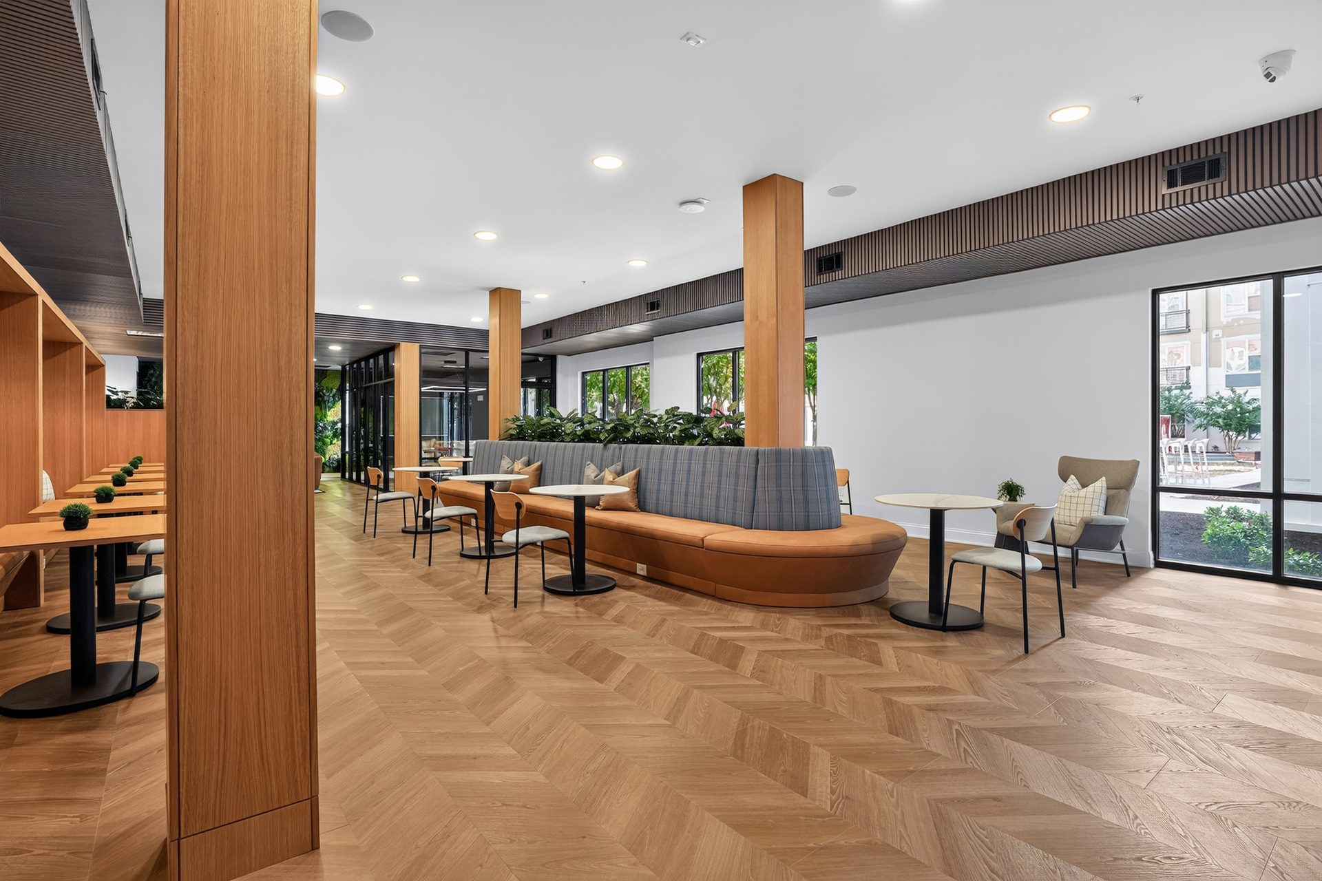 Open study lounge with a curved plaid banquette, wood columns, and windows onto the street