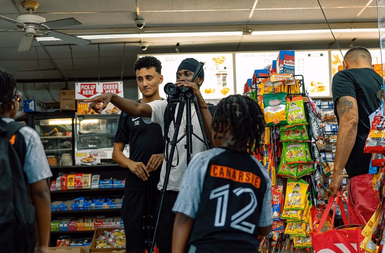 Two young men setting up a camera on a tripod inside a bodega with snacks shelves and customers around.