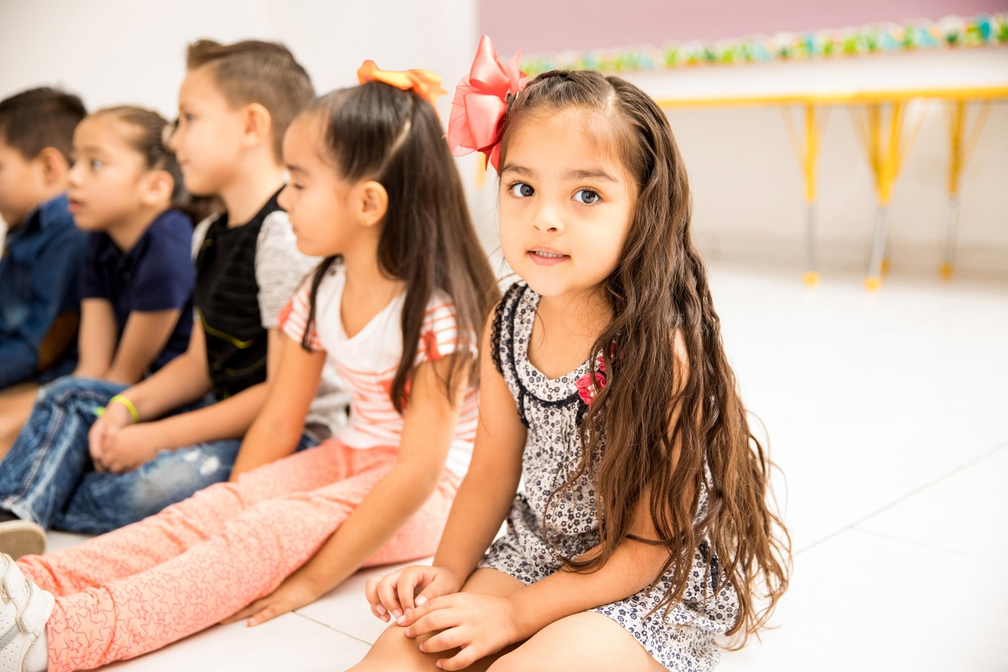 Five young children sitting on the floor in a row in a brightly lit classroom, with a girl in floral dress and pink bow in the foreground looking at the camera.