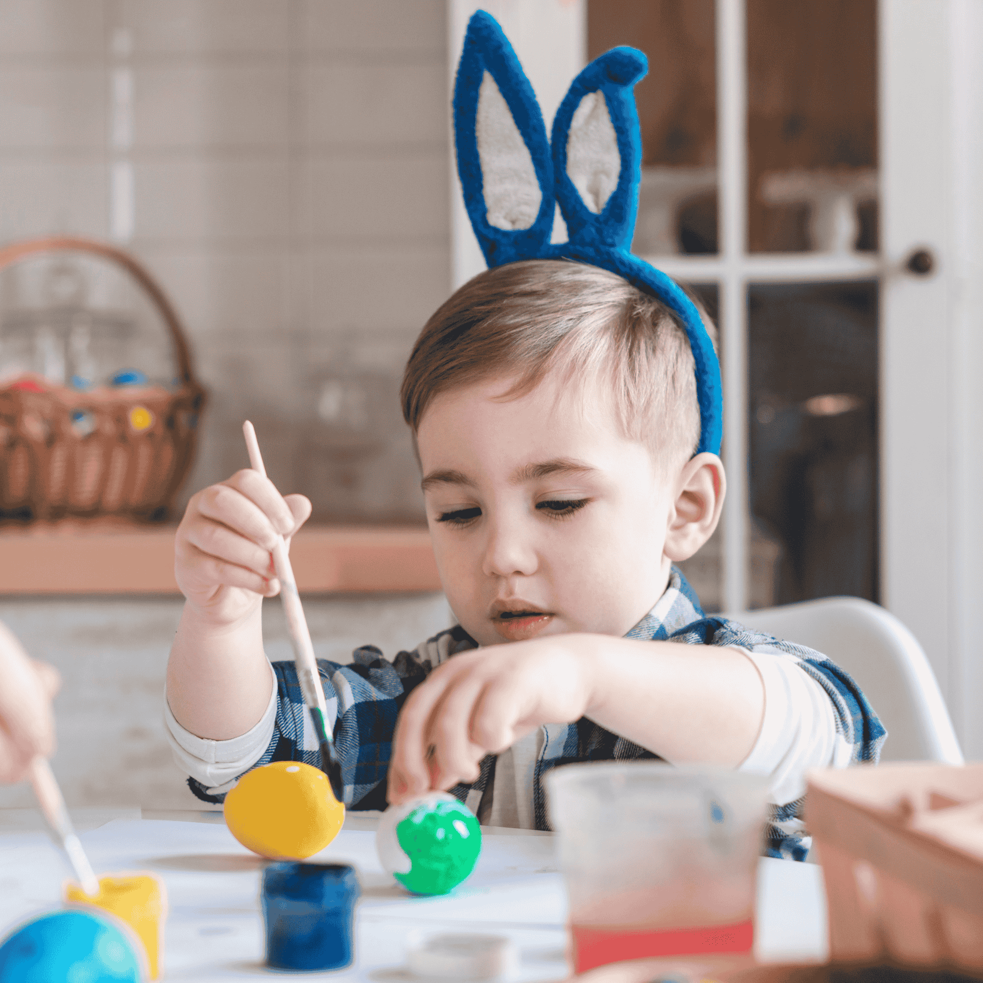 Young boy wearing blue bunny ears painting colorful Easter eggs at a table.