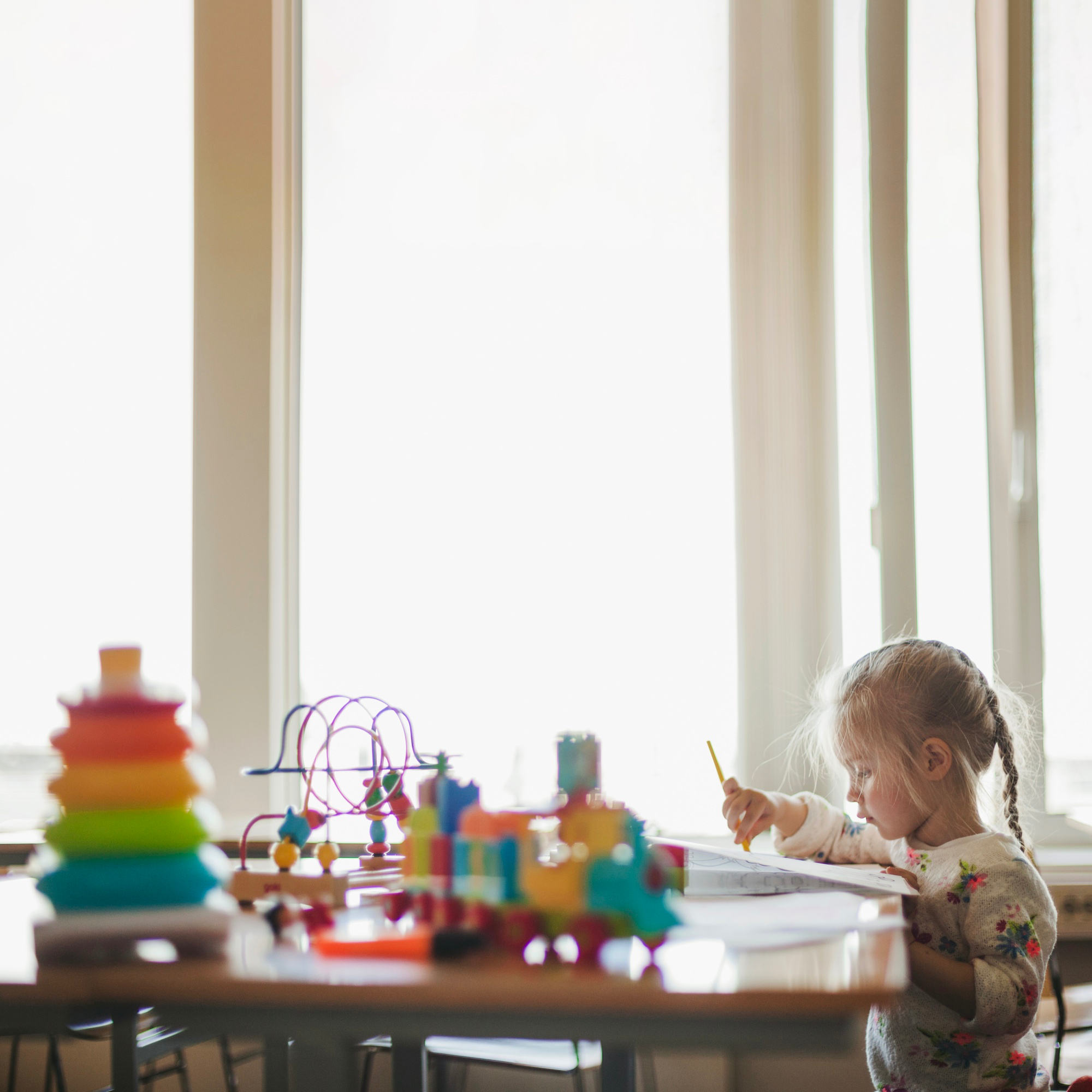 Young girl with a ponytail drawing with a yellow pencil at a table with colorful toys near a window.