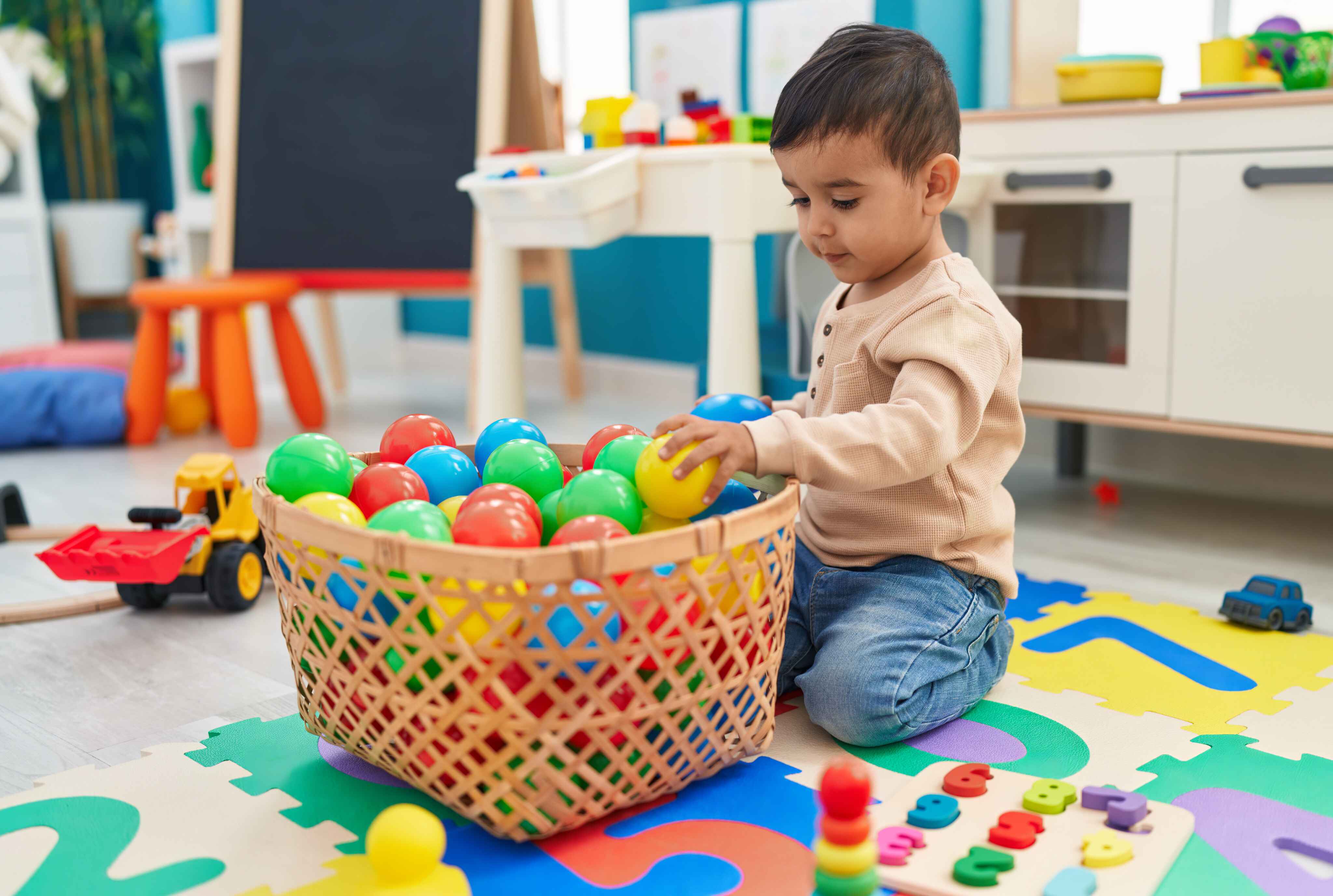Young child playing with colorful plastic balls in a basket on a foam alphabet puzzle mat.