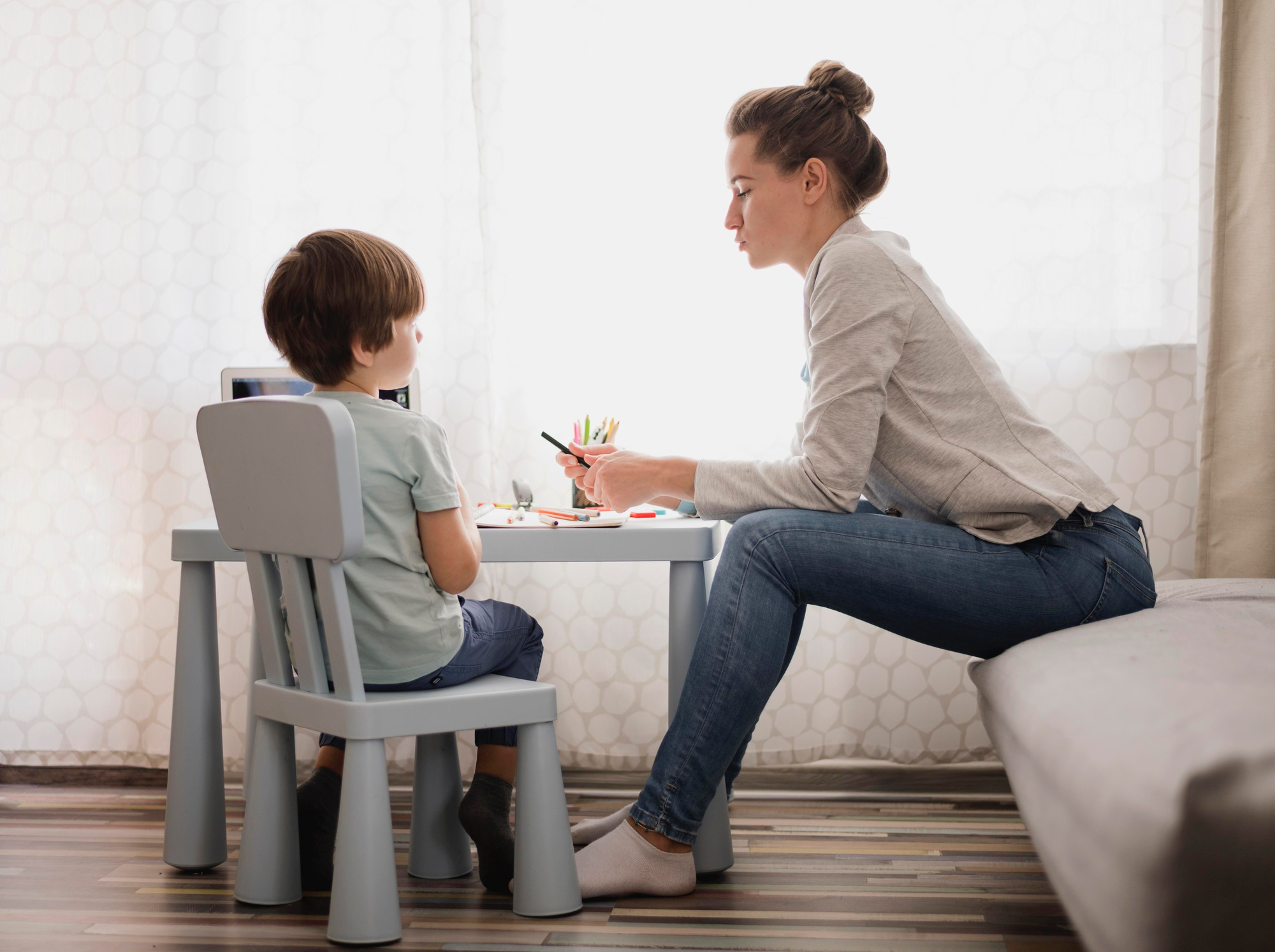 Woman sitting on a couch helping a young boy sitting at a small table with coloring utensils.