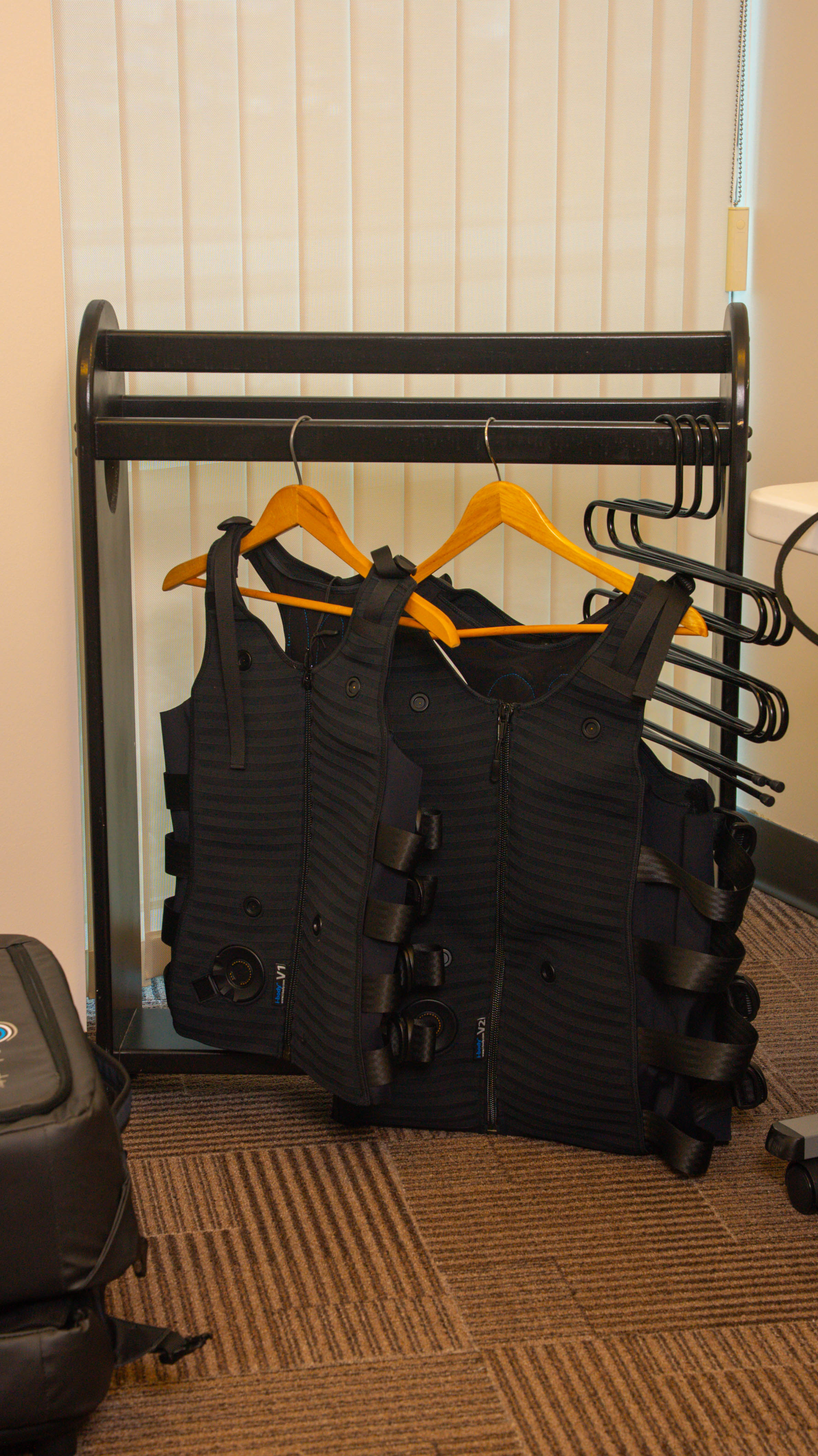Two black weighted vests hanging on wooden hangers on a black metal rack in a carpeted room.