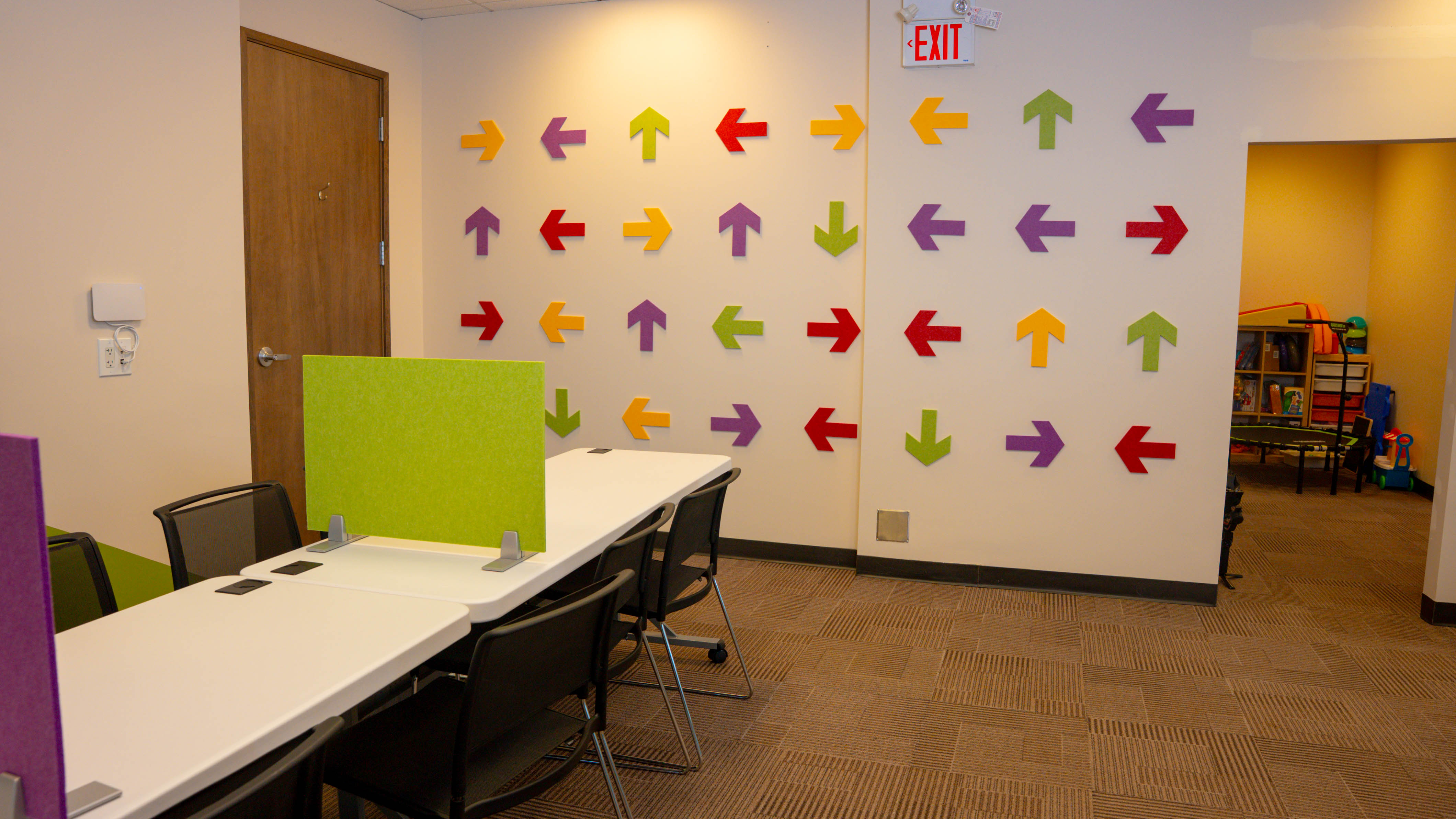 Office space with tables and chairs, colorful arrow decorations on the wall, and an exit sign above a doorway.