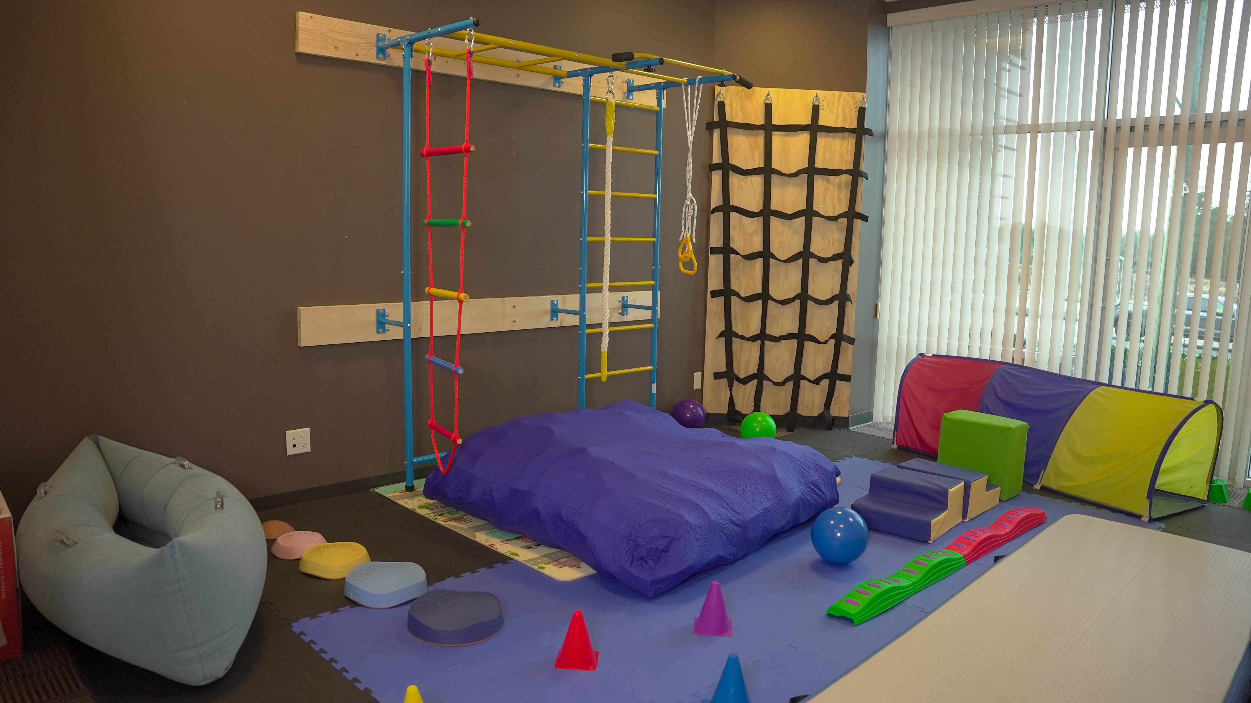 Indoor pediatric therapy room with colorful play and exercise equipment, including a climbing frame, soft mats, balls, cones, and a tunnel near a window with vertical blinds.