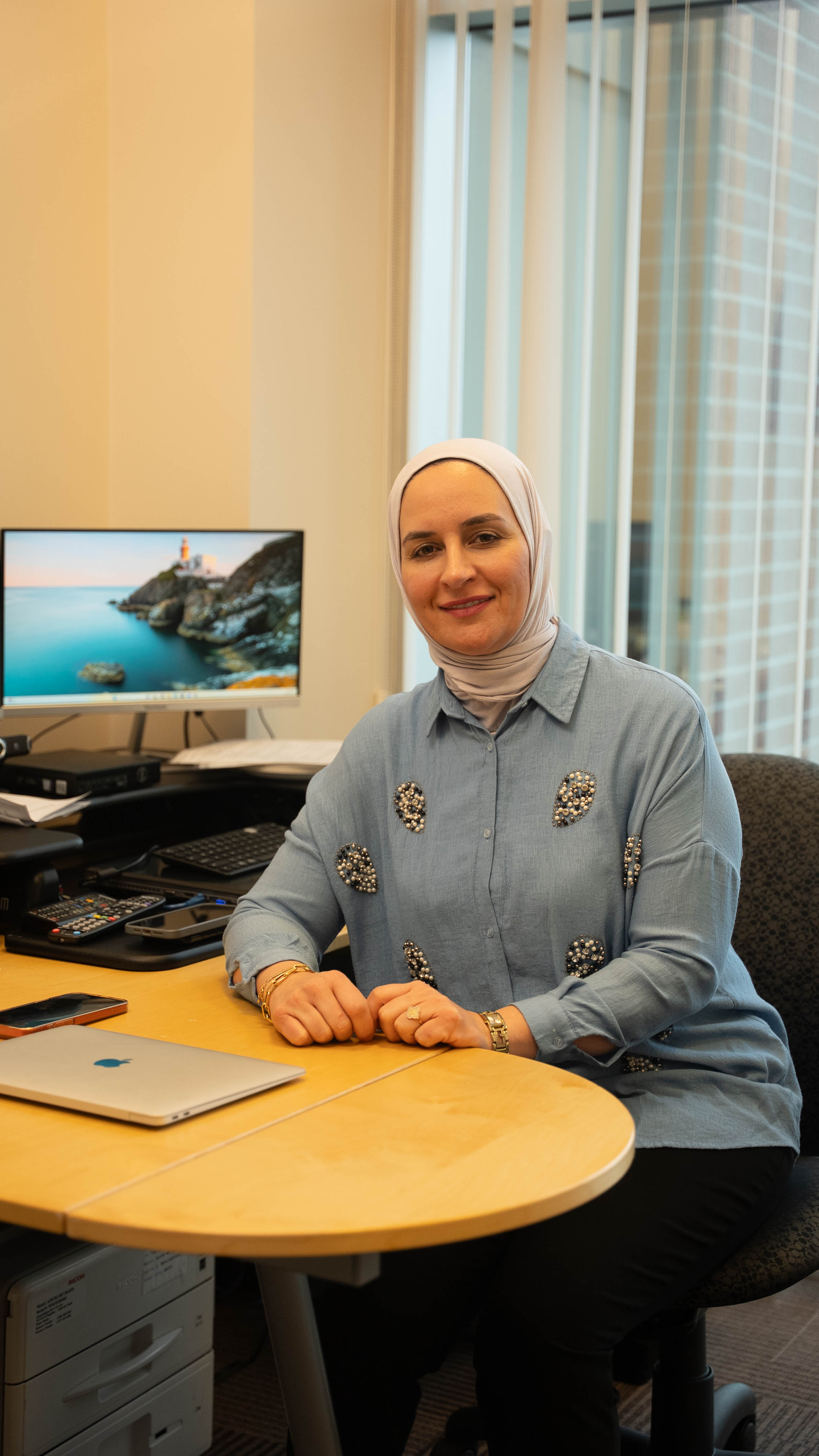 Woman wearing a light blue hijab and shirt with pearl embellishments sitting at an office desk with a laptop and monitor displaying a coastal scene.