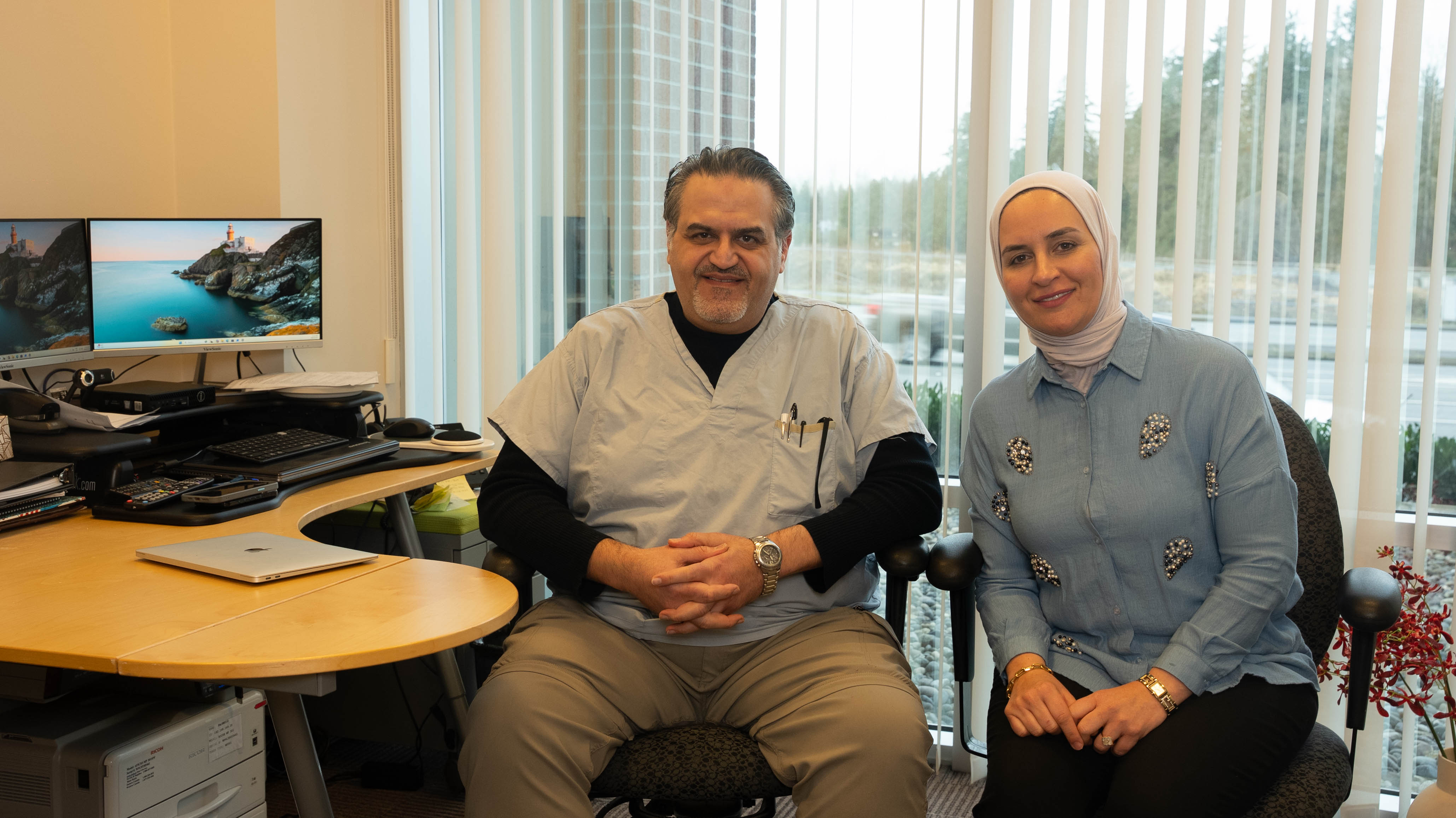 Two healthcare professionals sitting in an office with dual monitors and a large window behind them.