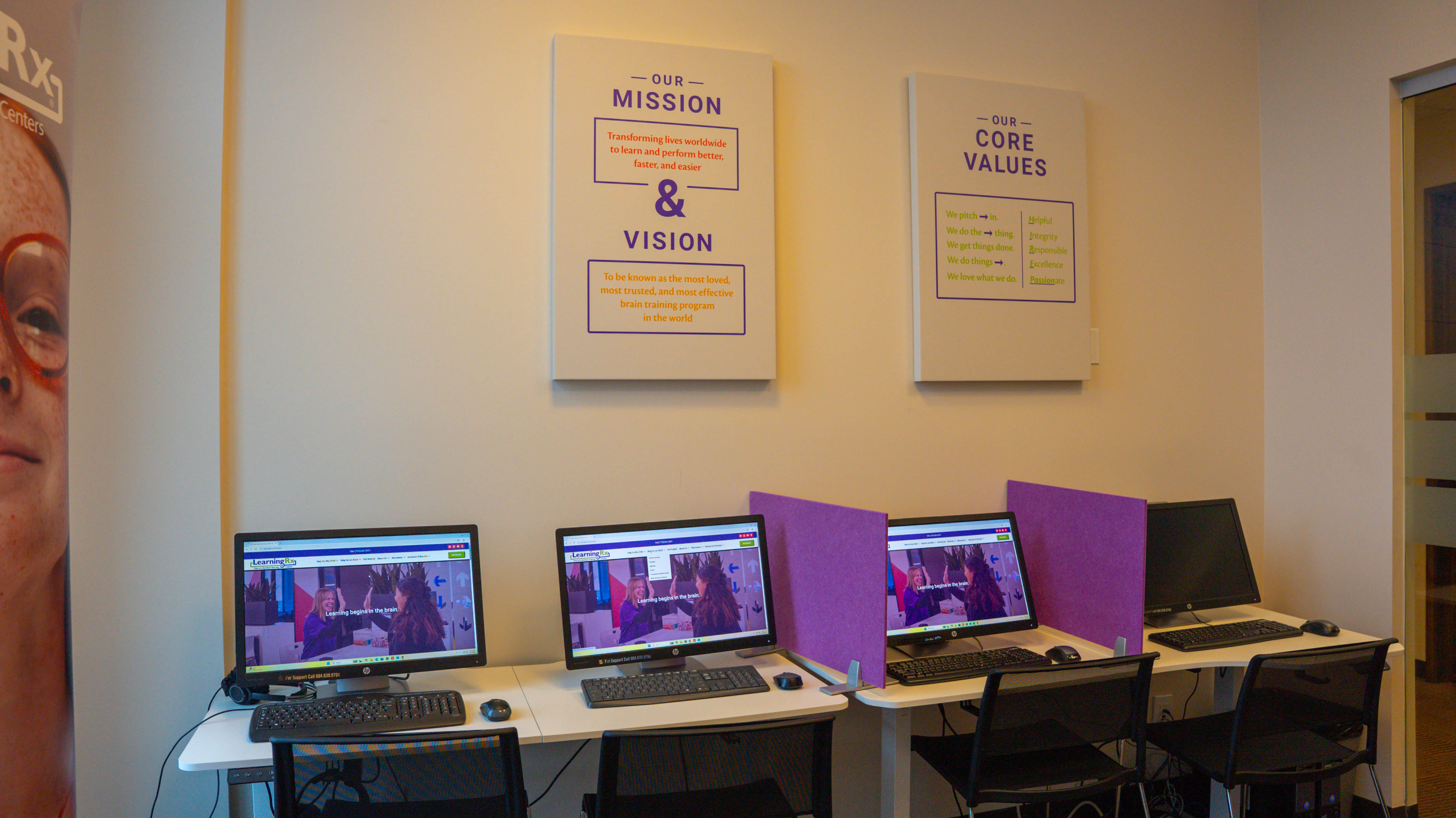 Four computer workstations with black chairs and keyboards in a room with white walls, purple privacy dividers, and wall signs displaying mission and core values.