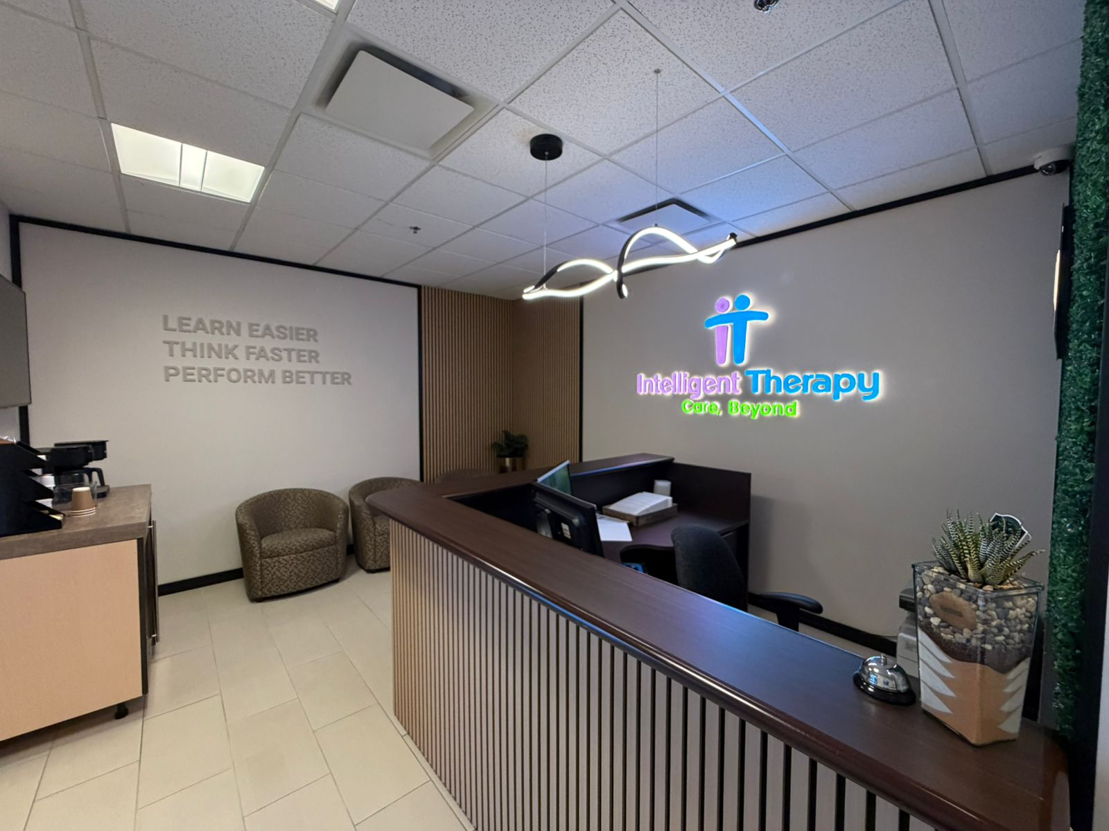 Reception area with a wooden desk, two armchairs, a coffee station, and wall signs reading 'Learn Easier Think Faster Perform Better' and 'Intelligent Therapy Care Beyond' with a logo.
