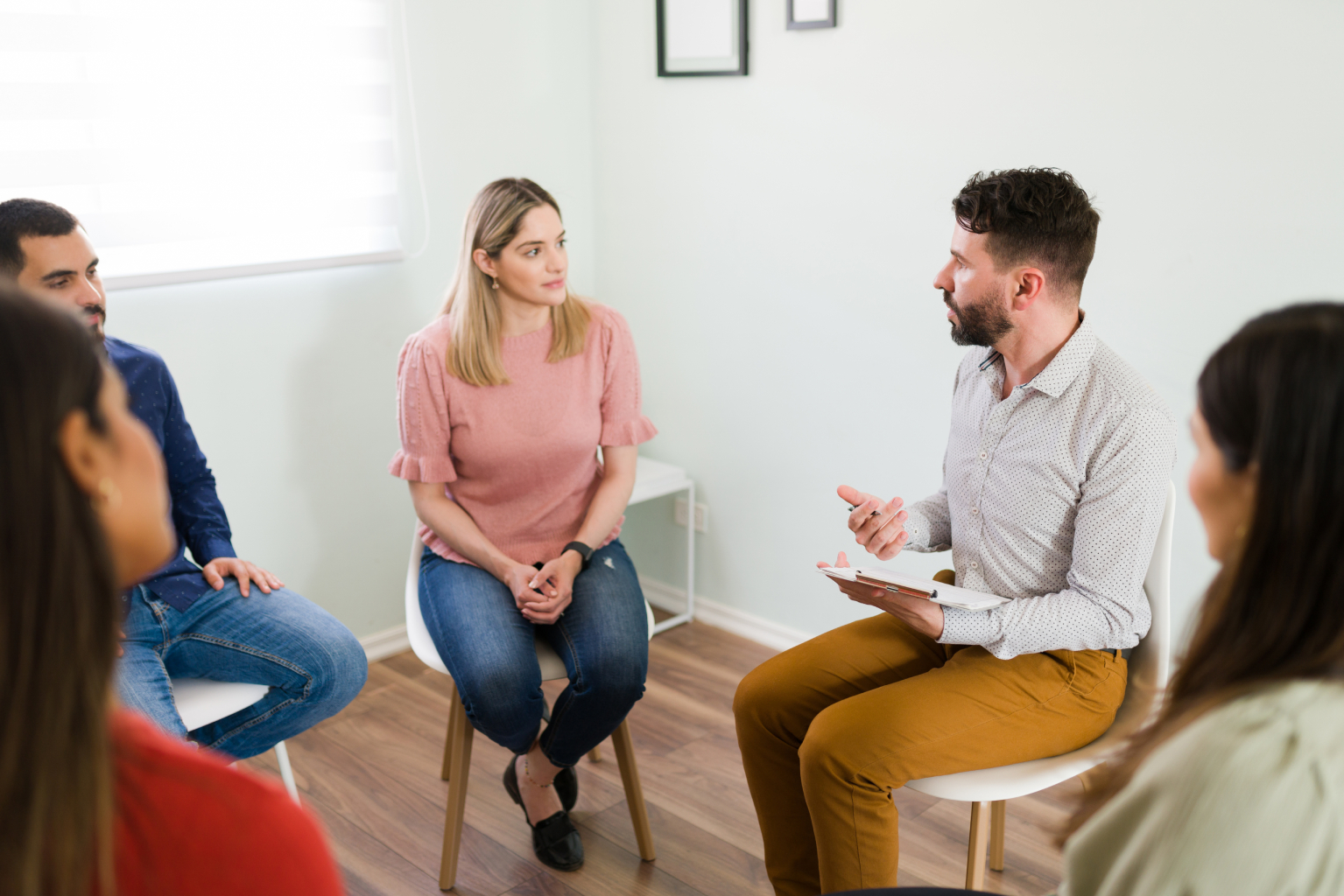 A group therapy session with a male therapist holding a clipboard talking to four seated participants in a bright room.